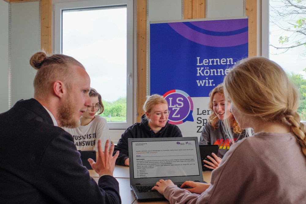 Einige Studierende sitzen am Tisch und lernen. Im Hintergrund sieht man das Logo des Lern- und Sprachenzentrums.