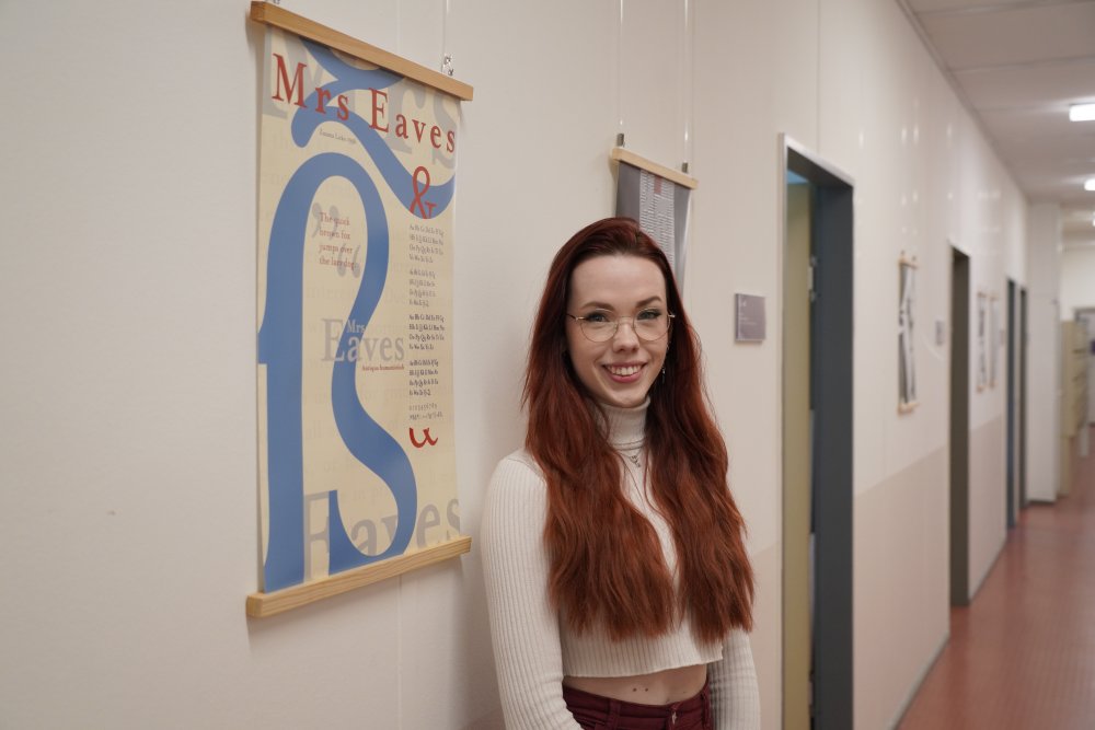 Eine junge Frau mit roten Haaren steht in einem Flur neben einem Plakat, auf dem ein großes ß abgebildet ist sowie der Schriftzug "Mrs Eaves" in verschiedenen Farben und weitere Schriftzüge. Im Hintergrund sieht man Türen und weitere Plakate.