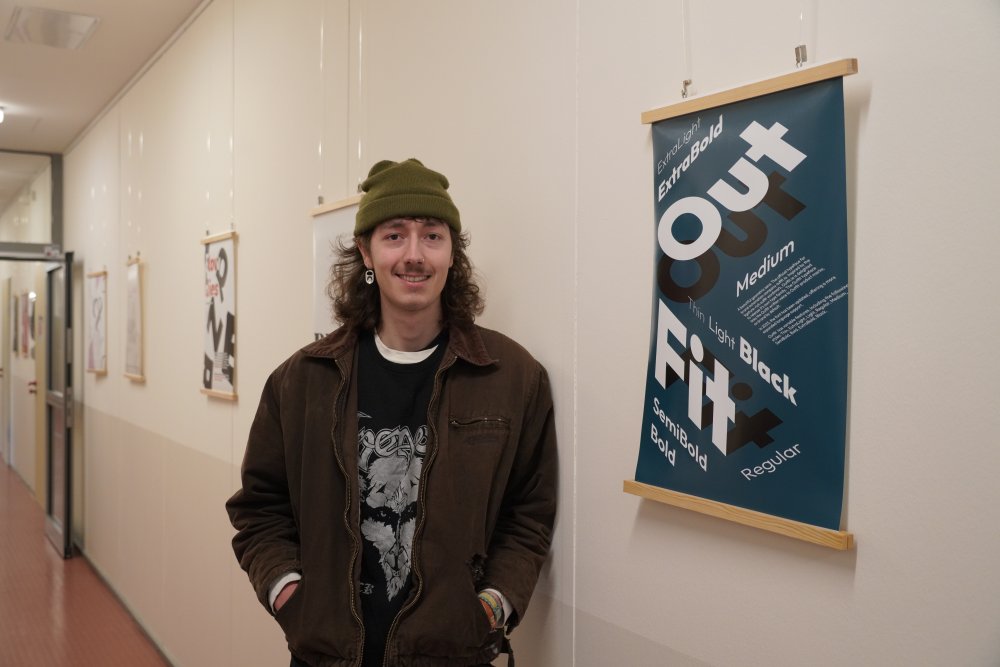 Ein junger Mann mit Wollmütze und Ohrring lehnt an einer Wand neben einem Plakat mit petrolfarbenem Hintergrund, dem Schriftzug "Out Fit", der Schatten schlägt, und weiteren Schriftzügen. Im Hintergrund sieht man weitere Plakate an der Wand hängen.