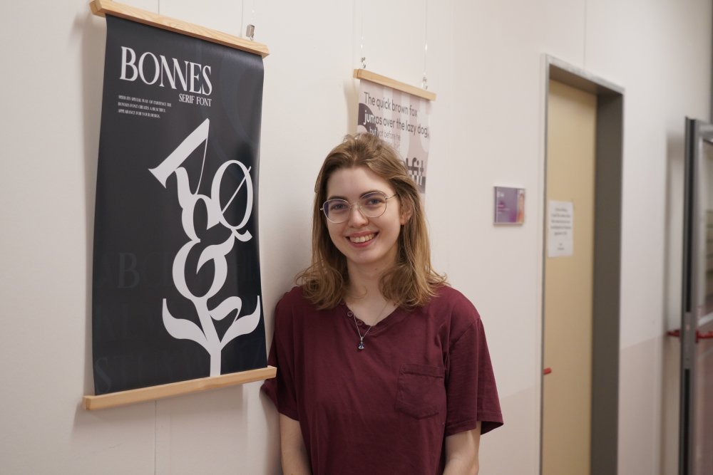 Eine junge Frau lehnt an einer Wand neben einem Plakat mit schwarzem Hintergrund, dem Schriftzug "Bonnes Serif Font" und einem Baum, der aus Schriftzeichen besteht. Im Hintergrund sieht man weitere Plakate an der Wand hängen.