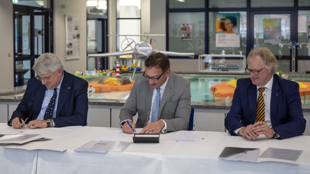 Drei Männer sitzen an einem Tisch, auf dem mehrere Dokumente ausgebreitet sind, zwei von ihnen setzen gerade ihre Unterschrift. Im Hintergrund sieht man ein Modell einer Hafenanlage.