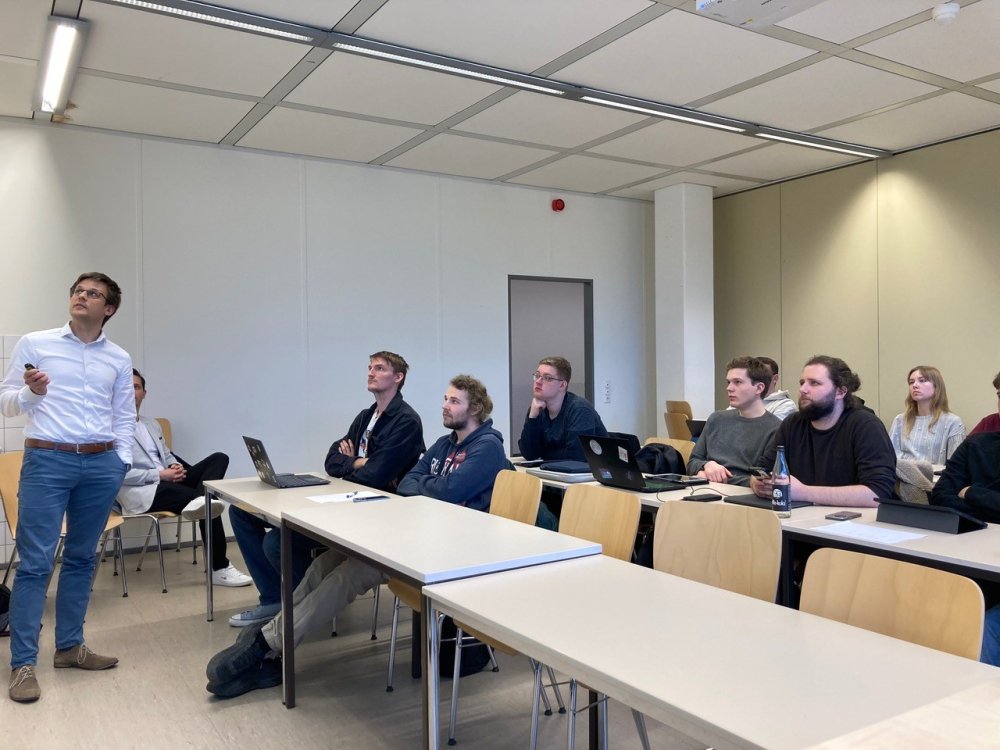Ein junger Mann im weißen Hemd steht vorne in einem Seminarraum und betätigt den Klicker in seiner Hand. An den Tischen sitzt eine Gruppe Studierender und schaut auf die Präsentation, die er gerade steuert.