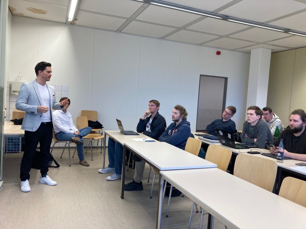 Ein junger Mann im weißen T-Shirt und hellgrauen Sportsakko steht vorne in einem Seminarraum und spricht. In dem Raum sitzt an den Tischen eine Gruppe Studierender.