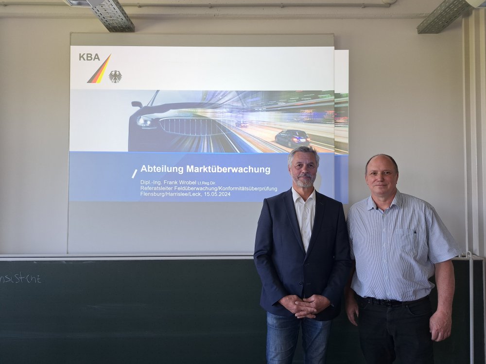 Zwei Männer, einer im Sakko und einer im blauen Hemd, stehen vor der Projektion einer Präsentations-Titelfolie, auf der zu lesen ist "Abteilung Marktüberwachung - Dipl.-Ing. Frank Wrobel, Referatsleiter Feldüberwachung/Konformitätsüberprüfung, Flensburg/Harrislee/Leck 15.05.2024". Dazu das Logo des KBA.