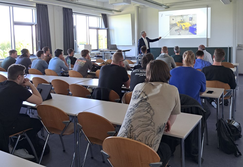 In einem Seminarraum sitzt eine Gruppe von Studierenden, vorne steht ein Mann im Sakko und zeigt mit einem Stab auf die Projektione einer Präsentationsfolie, auf der ein Auto zu sehen ist.