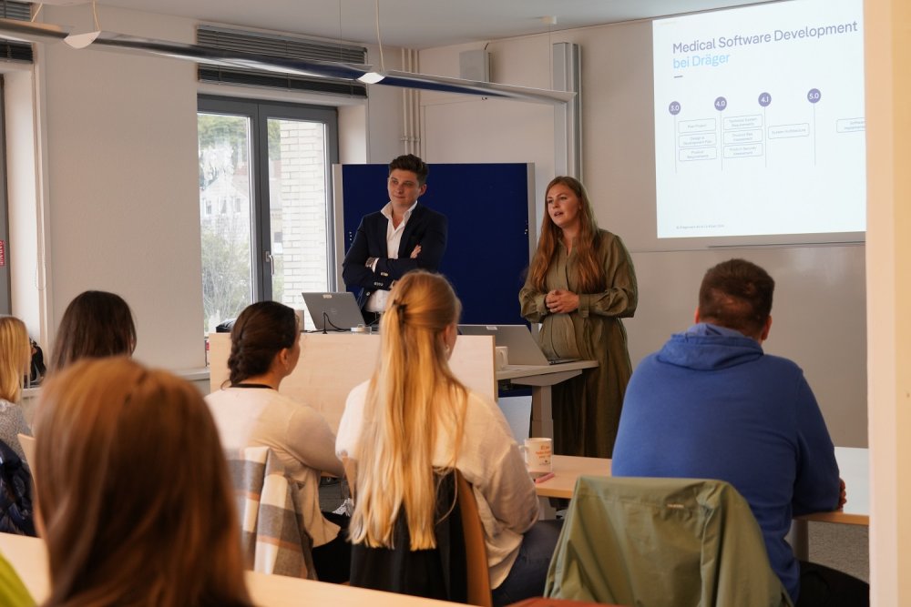 Ein Mann und eine Frau stehen vor einem Seminarraum mit Studierenden. Hinter ihnen an der Wand eine Präsentation mit der Überschrift "Medical Software Development bei Dräger".