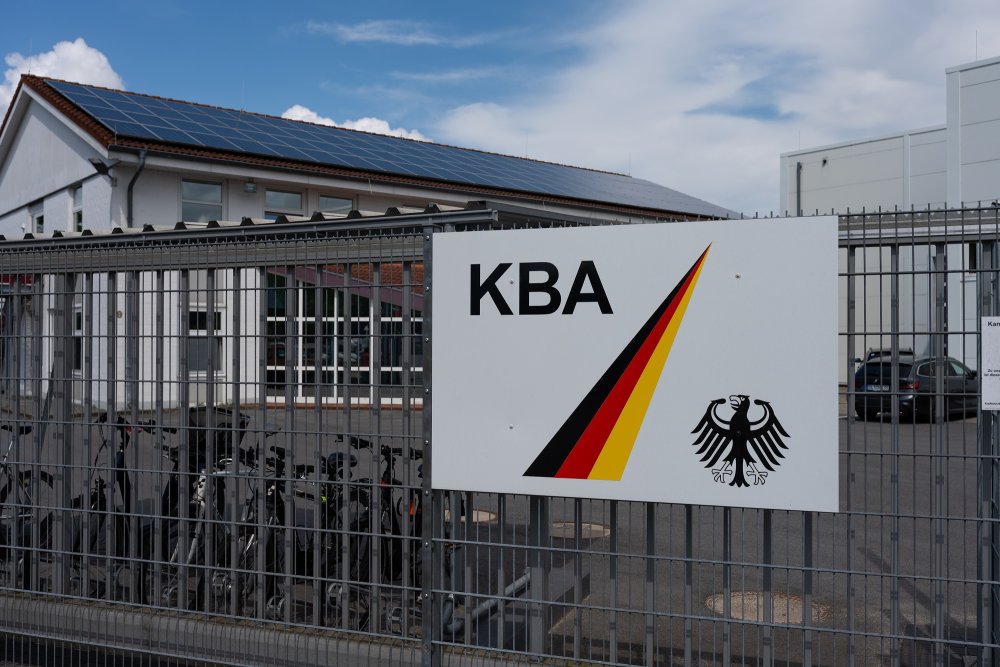 Ein Gebäude hinter einem Sicherheitszaun, an diesem hängt ein Schild mit den Buchstaben "KBA", einem Balken in schwarz, rot, gold und dem Bundesadler.