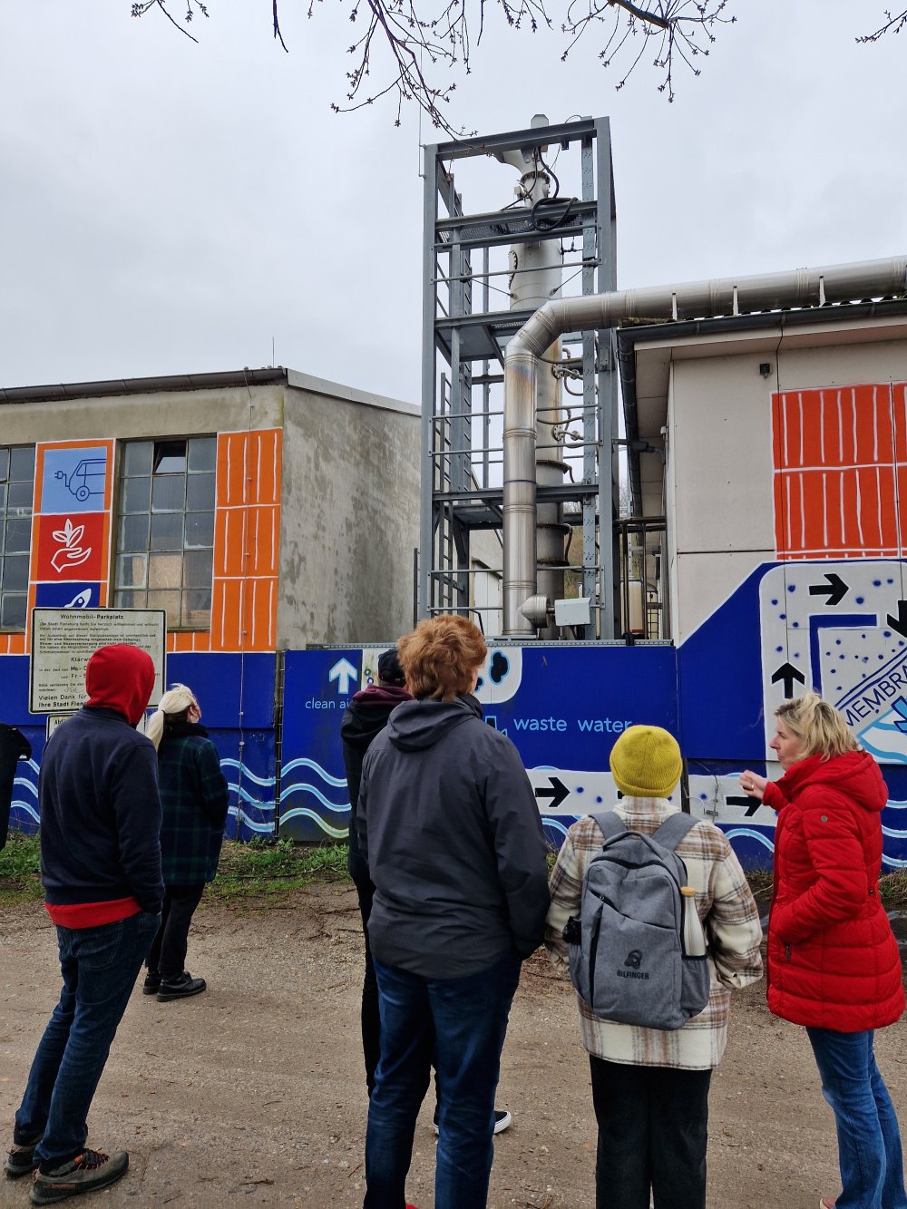 Eine Gruppe von Studierenden steht vor einem niedrigen, bunt bemalten Gebäude mit einem großen Abgasturm, während eine Dozentin ihnen etwas erklärt.