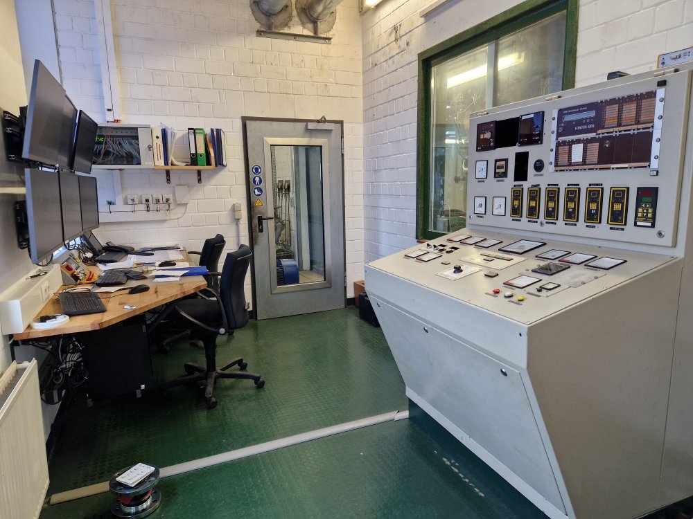 Blick in einen technischen Kontrollraum mit verschiedenen Schaltpanelen und einem Schreibtisch mit einem PC.