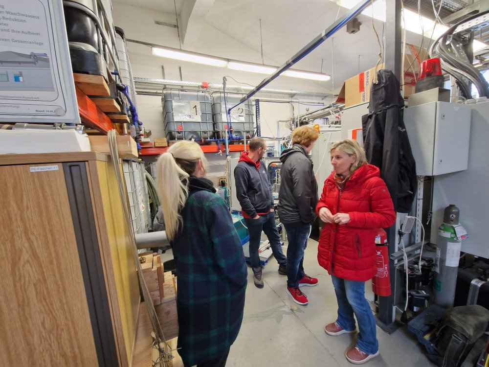 Eine Gruppe von Studierenden und eine Dozentin stehen in einem Lagerraum, durch den verschiedene Schläuche zu zwei großen Plastiktanks laufen.