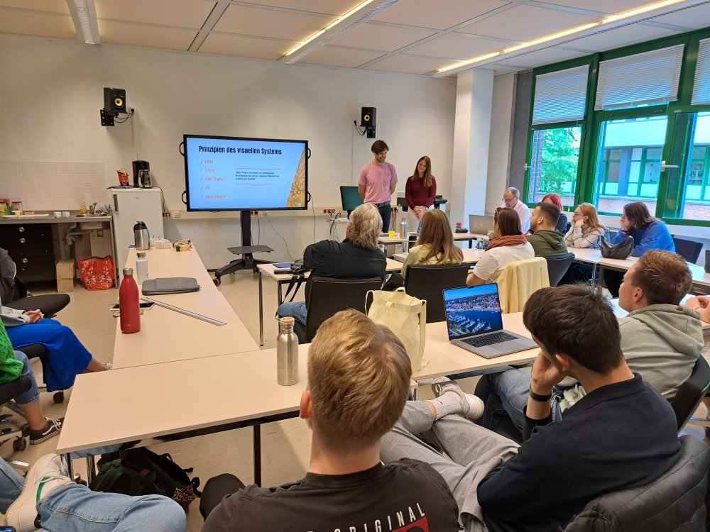 In einem Seminarraum stehen zwei Studierende vorn an einem großen Montor, auf dem eine Präsentationsfolie mit der Überschrift &quot;Prinzipien des visuellen Systems&quot; zu sehen ist.
