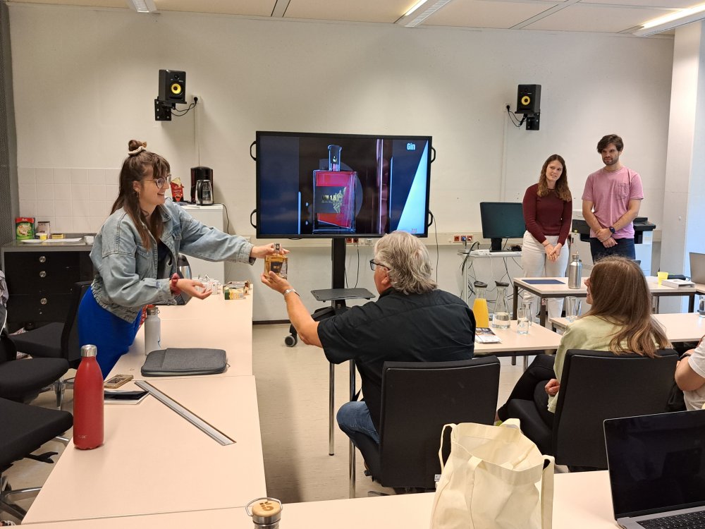 In einem Seminarraum stehen zwei Studierende vorne an einem großen Monitor, während eine andere Studierende im Raum einem Mann eine Glasflasche reicht.