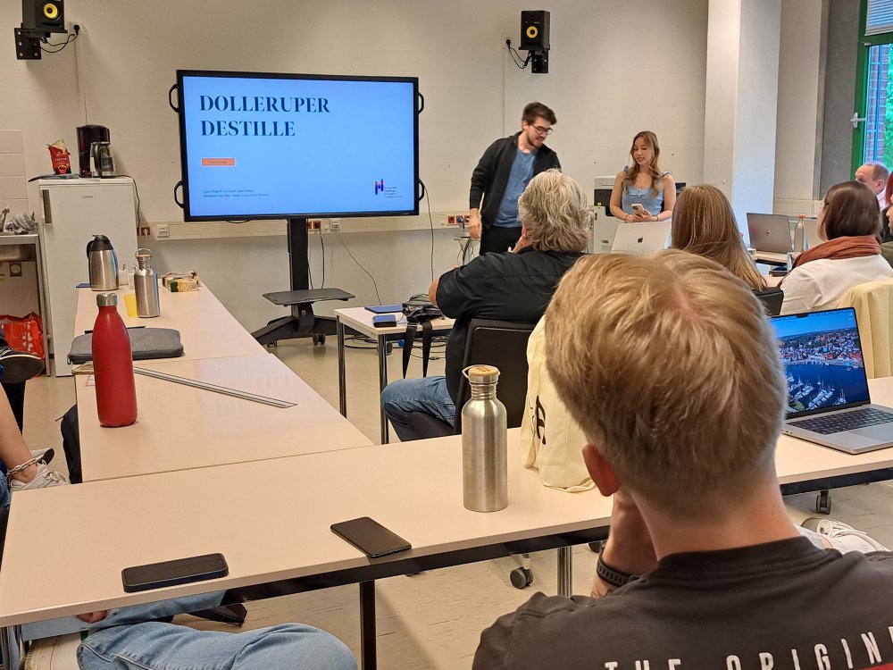 In einem Seminarraum stehen zwei Studierende vorne an einem großen Monitor und referieren, weitere Studiernde sitzen und hören zu.