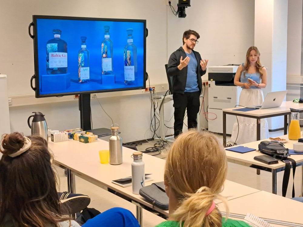 In einem Seminarraum stehen zwei Studierende vor einem großen Monitor, auf dem ein Hochglanz-Werbefoto von vier Schnapsflaschen zu sehen ist.