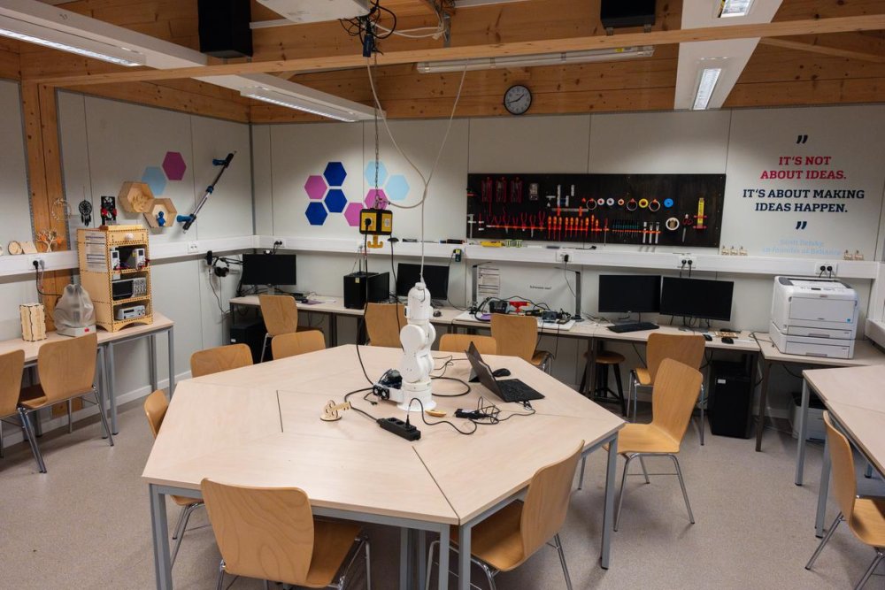 In einem Raum steht ein sechseckiger Tisch mit Stühlen, darauf ein weißer Roboterarm. An den Wänden weitere Tische und Stühle mit Monitoren und Werkzeug, an den Wänden bunte Sechsecke und Schrift.