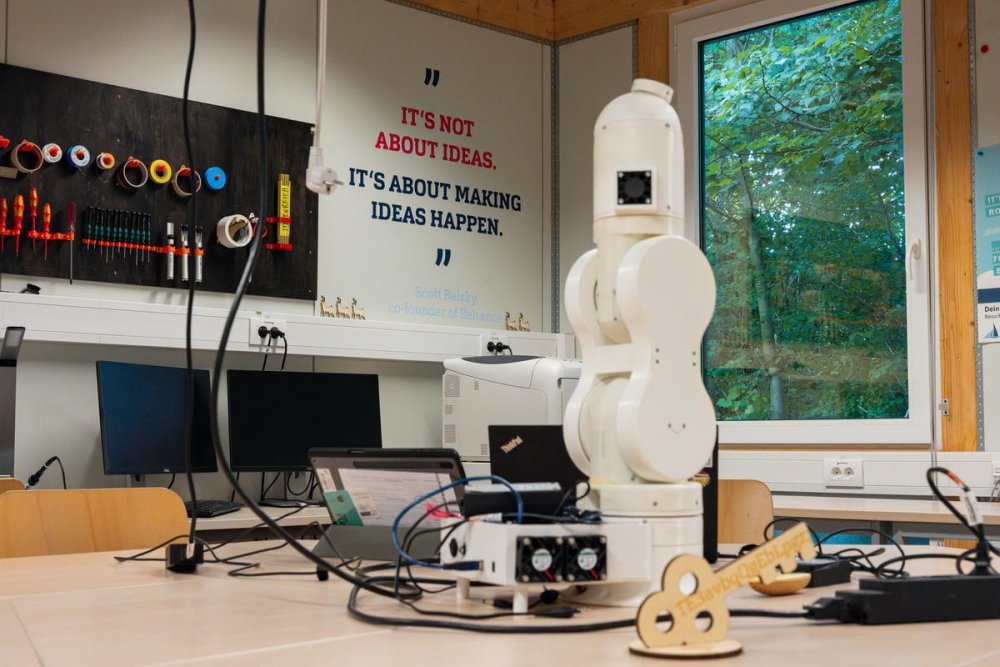 Auf einem Tisch steht ein weißer Roboterarm, an der Wand dahinter steht in bunter Schrift: "It's not about ideas, it's about making ideas happen".