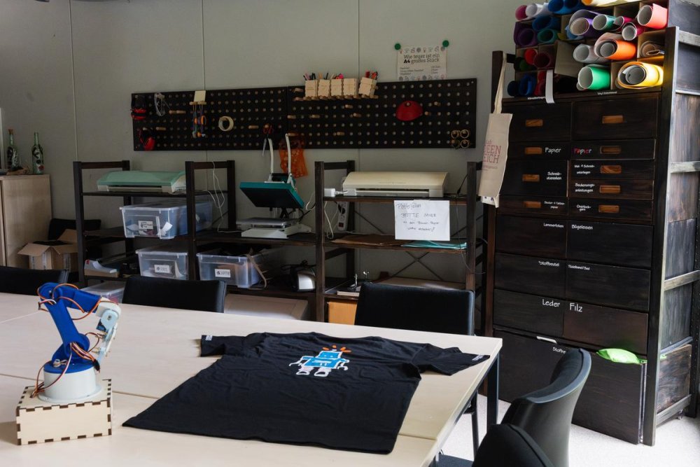 In einem Raum steht ein Tisch, darauf ist ein schwarzes T-Shirt mit dem Aufdruck eines gezeichneten Roboters und ein kleiner Roboterarm. An der Wand dahinter Regale mit bunten Papierrollen, Plastikboxen und Schubladen mit Aufschriften wie Leder, Filz, Papier und anderen.