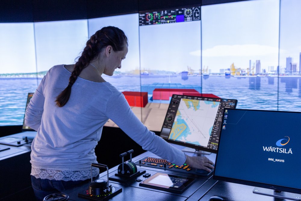 Eine junge Frau steht in einem Schiffsführungssimulator, im Hintergrund sieht man die Projektion eines Handelshafens.