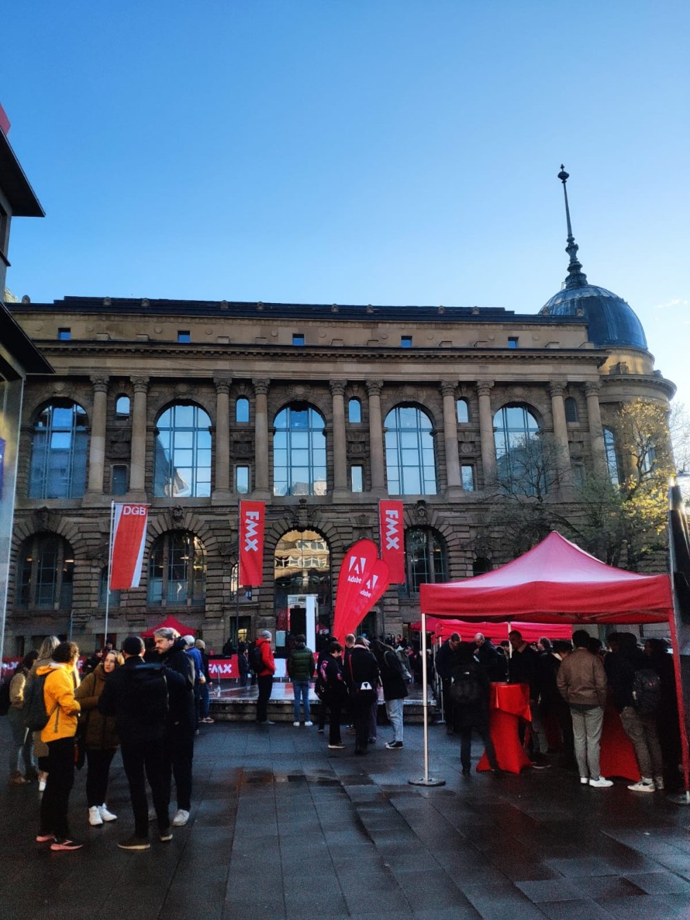 Ein großes Sandsteingebäude, an dem rote Flaggen mit der Aufschrift "FMX" hängen, davor ein Platz mit ebenfalls roten Pavillons und Beachflags.