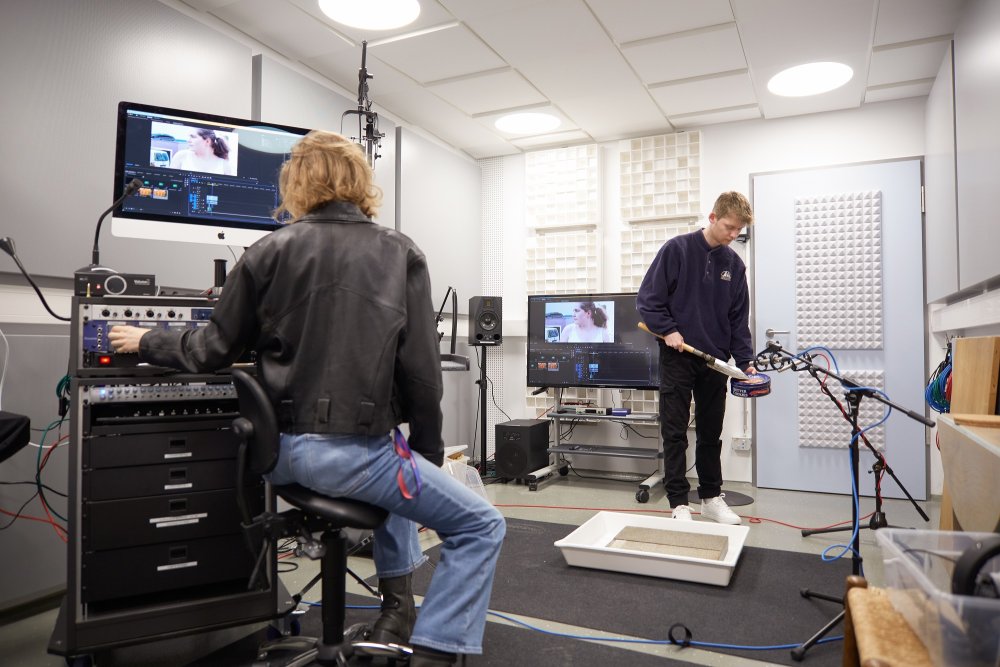In einem Studioraum sitzt eine junge Frau in schwarzer Lederjacke an einem Mischpult, über dem ein Monitor aufgebaut ist, an dem ein Videobearbeitungsprogramm zu sehen ist. Ein junger Mann kniet vor einem Mikrofon und reibt mit einem Metallspaten über eine Keksdose.
