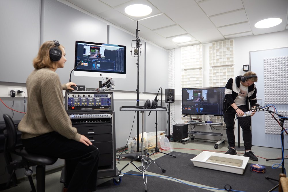 In einem Studioraum sitzt eine junge Frau an einem Mischpult, über dem ein Monitor aufgebaut ist, an dem ein Videobearbeitungsprogramm zu sehen ist. Ein junger Mann steht vor einem Mikrofon und öffnet eine Plastikflasche.