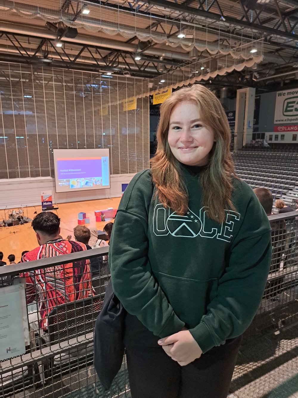 Eine junge Frau im grünen Pullover steht vor einer Tribüne, im Hintergrund sieht man eine Bühne mit einer Leinwand, auf die eine Präsentation projeziert wird.