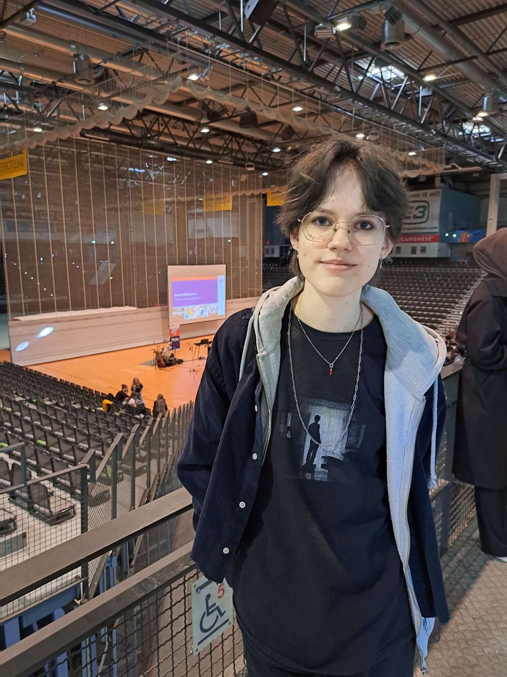 Eine junge Frau in schwarzer Kapuzenjacke steht vor einer Tribüne, im Hintergrund sieht man eine Bühne mit einer Leinwand, auf die eine Präsentation projeziert wird.