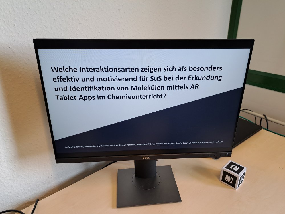 Auf einem Schreibtisch steht ein Monitor, auf dem eine Präsentation mit Text zu sehen ist: "Welche Interaktionsarten zeigen sich als besonders effektiv und motivierend für SuS bei der Erkundung und identifikation von Molekülen mittels AR Tablet-Apps im Chemieunterricht?"