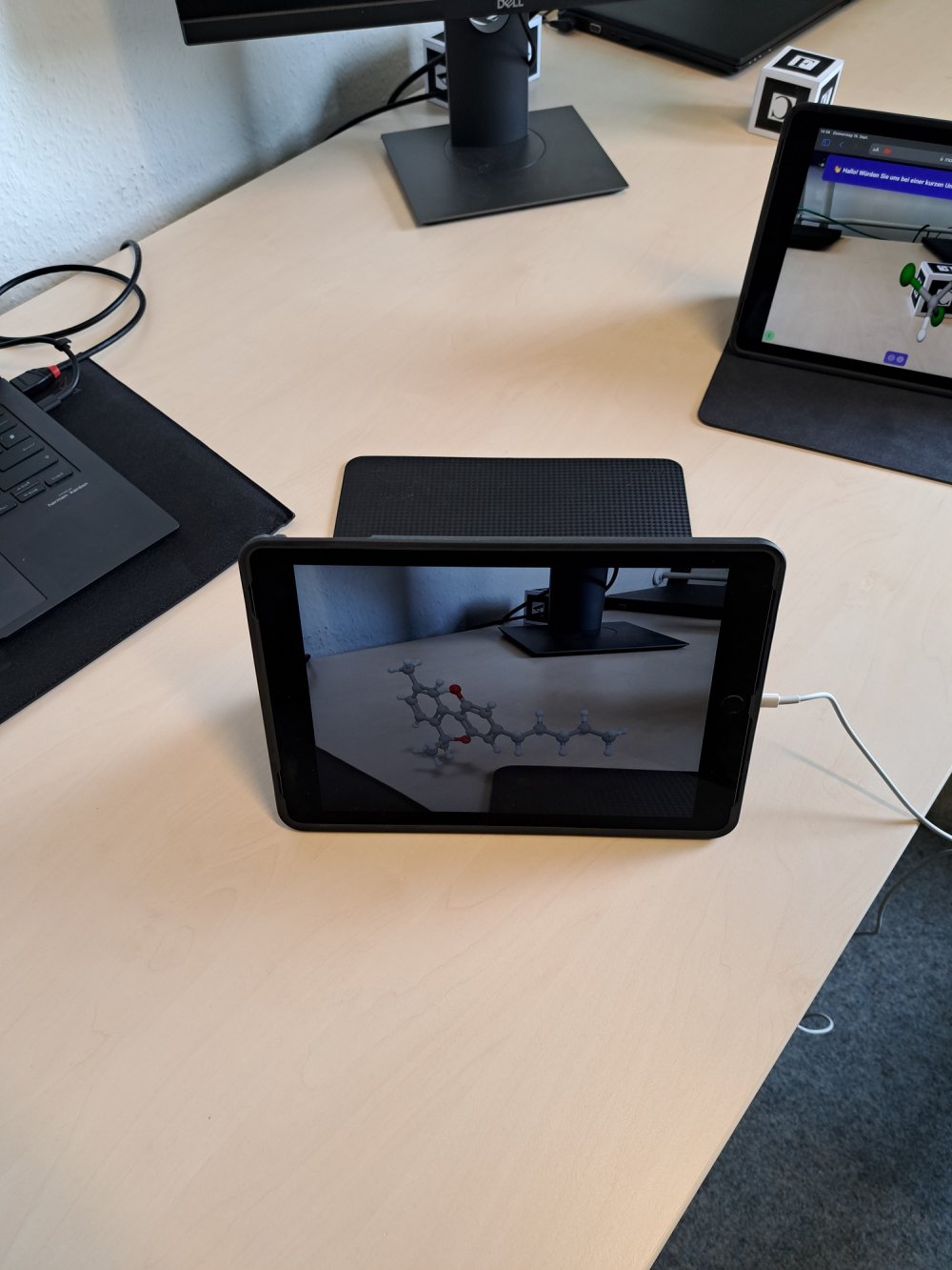 Auf einem Schreibtisch steht ein Tablet, auf dem ein virtuelles Modell eines Moleküls zu sehen ist.