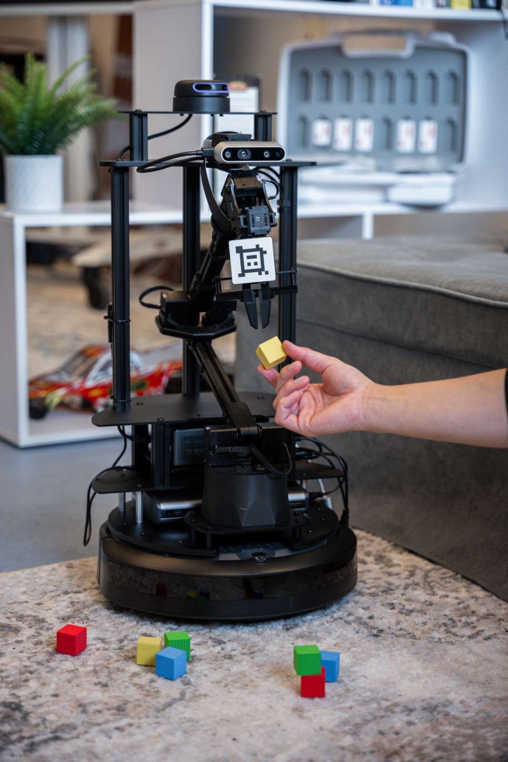 Auf einem Teppich steht ein Roboterarm, eine Hand reicht ihm einen bunten Bauklotz, vor ihm liegen weitere auf dem Boden.