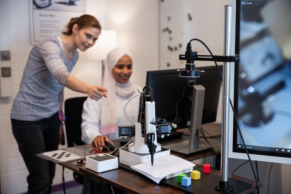 Eine junge Frau mit Kopftuch sitzt an einem PC-Arbeitsplatz, eine andere Frau steht neben ihr und erklärt ihr etwas am Bildschirm. Im Vordergrund steht ein Roboterarm.