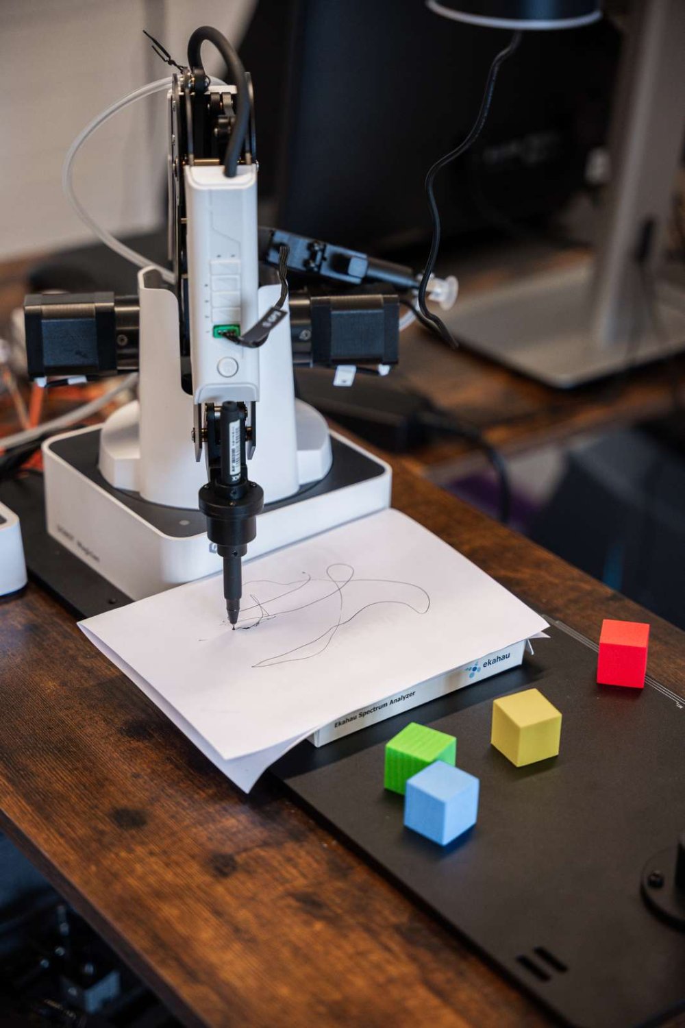 Auf einem Schreibtisch steht ein Roboterarm, der mit einem Stift auf Papier malt.