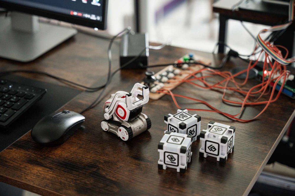 Auf einem Schreibtisch steht ein kleiner Roboter vor drei Kunststoff-Würfeln, dahinter sieht man eine Schaltvorrichtung mit vielen bunten Kabeln und die Ecke eines PC-Monitors.