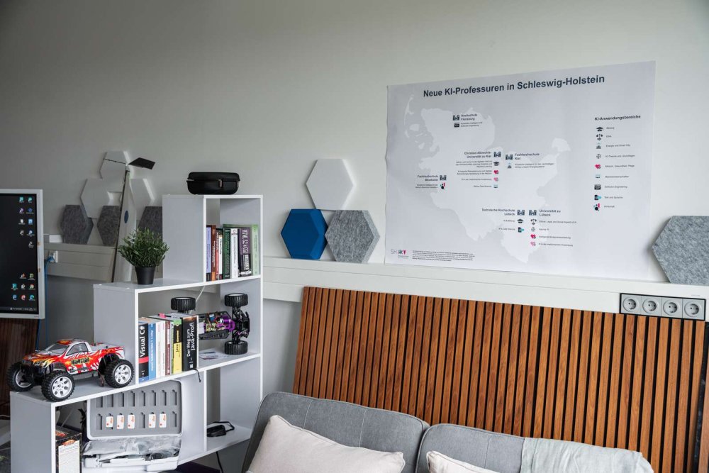 In einem Raum steht ein Sofa und ein Regal, an der Wand darüber hängt eine Karte mit der Überschrift &quot;Neue KI-Professuren in Schleswig-Holstein&quot;.