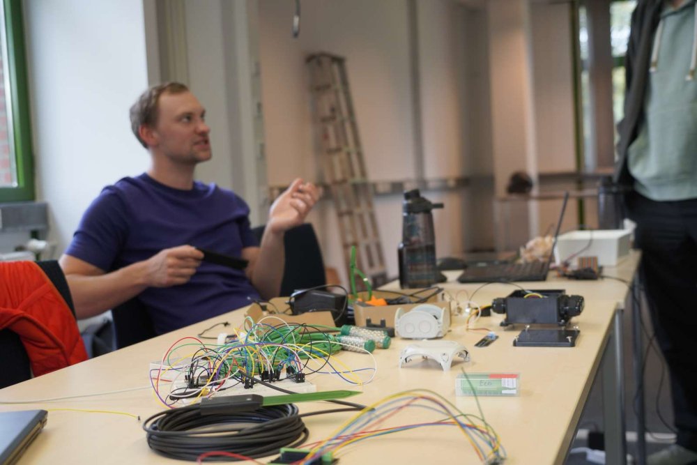 An einem Tisch sitzt ein Student vor einer Menge an miteinander verkabelten Sensoren und spricht mit einer Person, die vor dem Tisch steht.