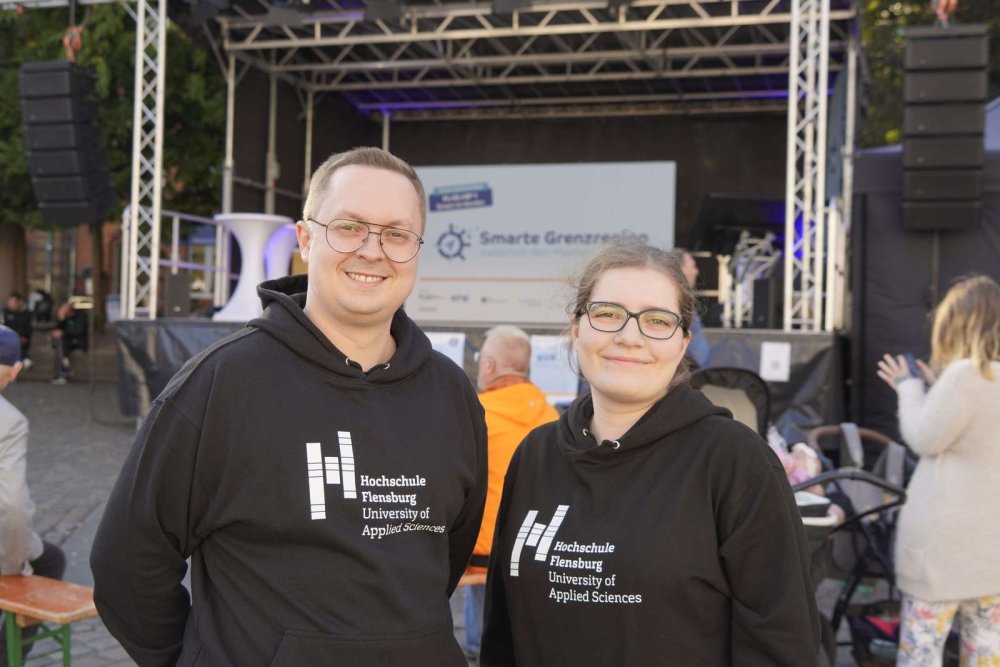Vor einer Bühne stehen eine Studentin und ein Student in schwarzen Kapuzenpullis mit dem Logo der Hochschule Flensburg.