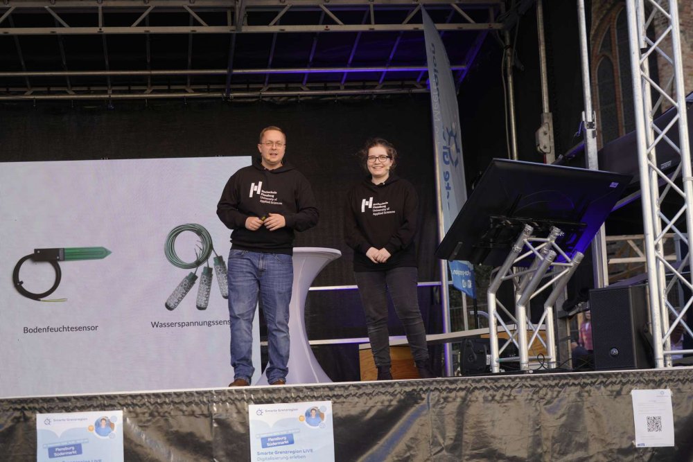 Zwei Studierende in schwarzen Kapuzenpullis mit dem Logo der Hochschule Flensburg stehen auf einer Bühne, hinter ihnen wird eine Präsentation an die Leinwand projeziert, auf der zwei Geräte mit den Unterschriften "Bodenfeuchtesensor" und "Wasserspannungssensor" zu sehen sind.