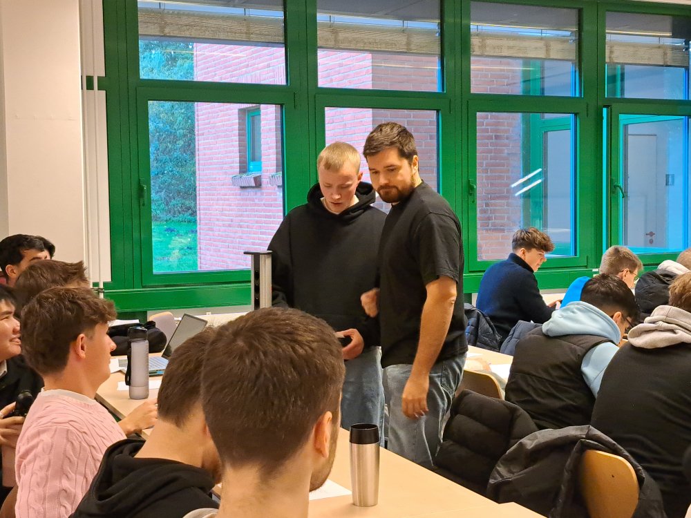 In einem Seminarraum stehen zwei Studierende zwischen zwei Tischreihen, die ebenfalls mit Studierenden und Schüler*innen voll besetzt sind, und sprechen mit den Schülern vor sich.