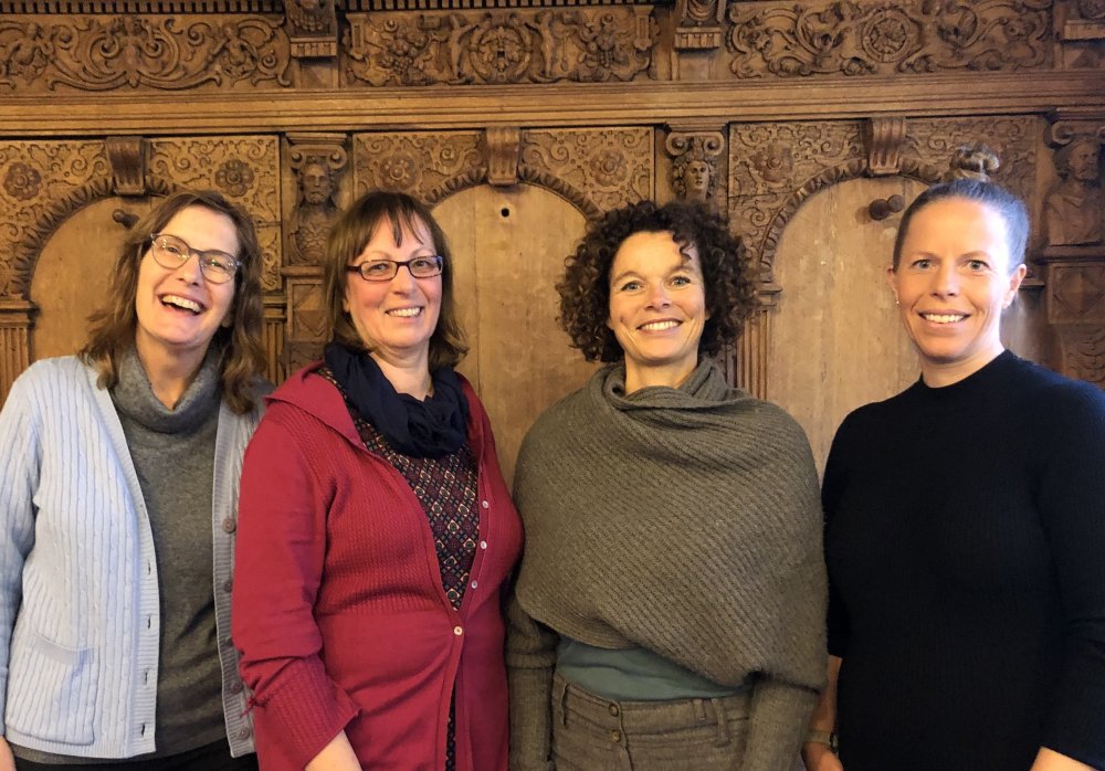 Vier Frauen stehen vor einer mit kunstvollen Schnitzereien verzierten Holzwand und lächeln offen in die Kamera. Sie tragen (von links nach rechts) einen blauen Cardigan mit Rollkragenpullover, einen roten Cardigan mit geblümter Bluse, einen braunen Wollpullover und ein schwarzes Landarmshirt.