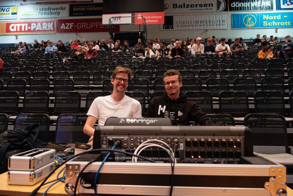 Zwei junge Männer sitzen in der ersten Reihe einer Tribüne, vor ihnen ein Mischpult.