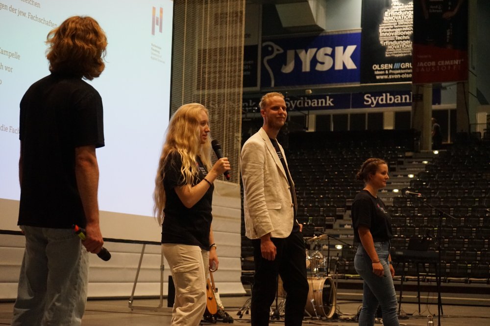 Vier Studierende stehen auf einer Bühne, eine von ihnen spricht gerade in ein Mikrofon, hinter ihnen sieht man angeschnitten eine Leinwand.