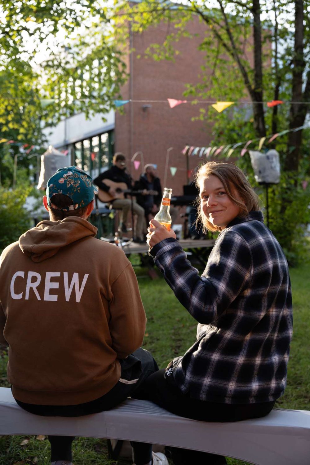 Eine junge Frau und ein junger Mann sitzen auf einer Bierbank in einem Garten, im Hintergrund ein großes Backsteingebäude. Die junge Frau dreht sich zum Betrachter um und prostet mit einer Limo-Flasche zu, der junge Mann trägt einen Pulli, auf dem in Großbuchstaben "Crew" aufgedruckt ist.