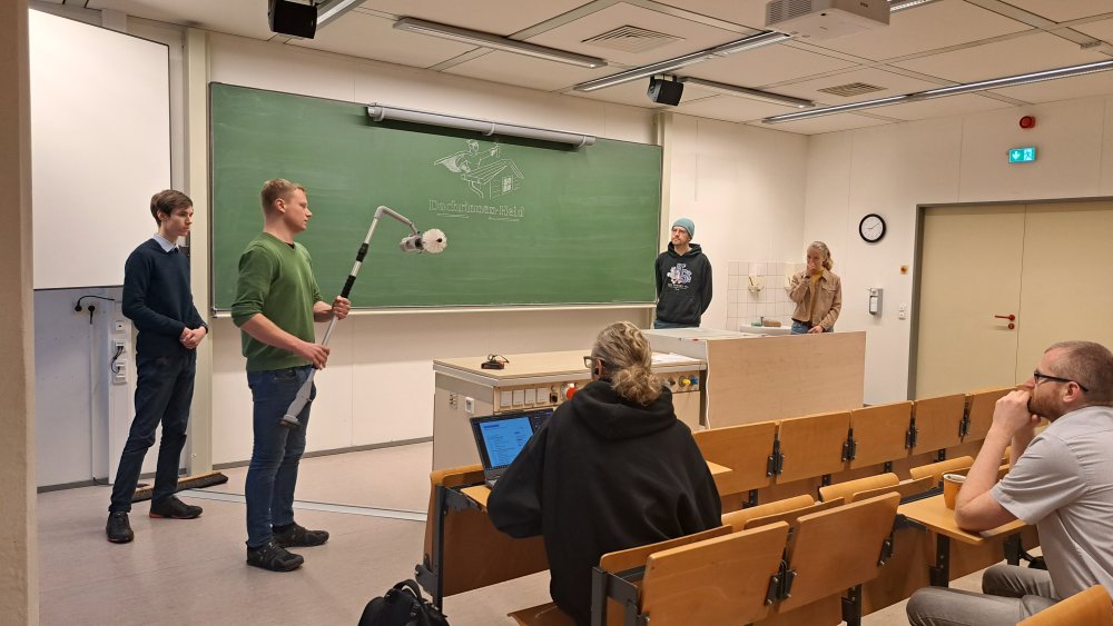 Eine Gruppe von Studierenden steht vorne in einem Vorlesungssaal und hält eine Präsentation. Einer der Studierenden hält ein elekrtisches Gerät mit Teleskoparm in der Hand, an der Tafel hinter ihnen ist ein Logo angezeichnet mit Schriftzug "Dachrinnen-Held"