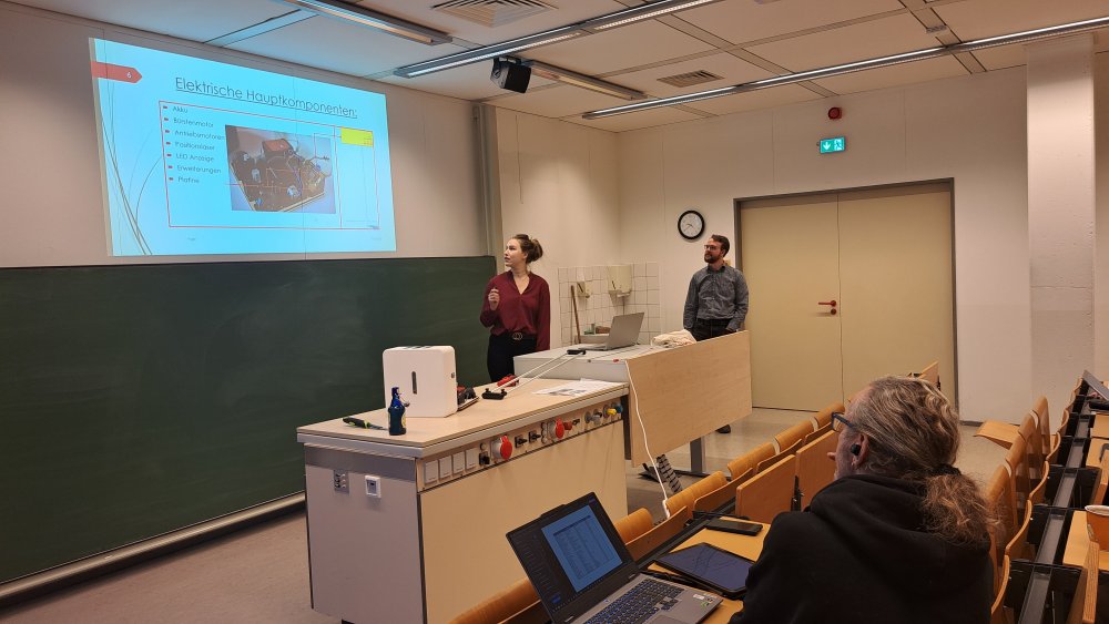 In einem Seminarraum steht eine Gruppe von Studierenden vorne und hält eine Präsentation. In der Präsentation sieht man ein Foto mit der Bildüberschrift "Elektrische Hauptkomponenten"