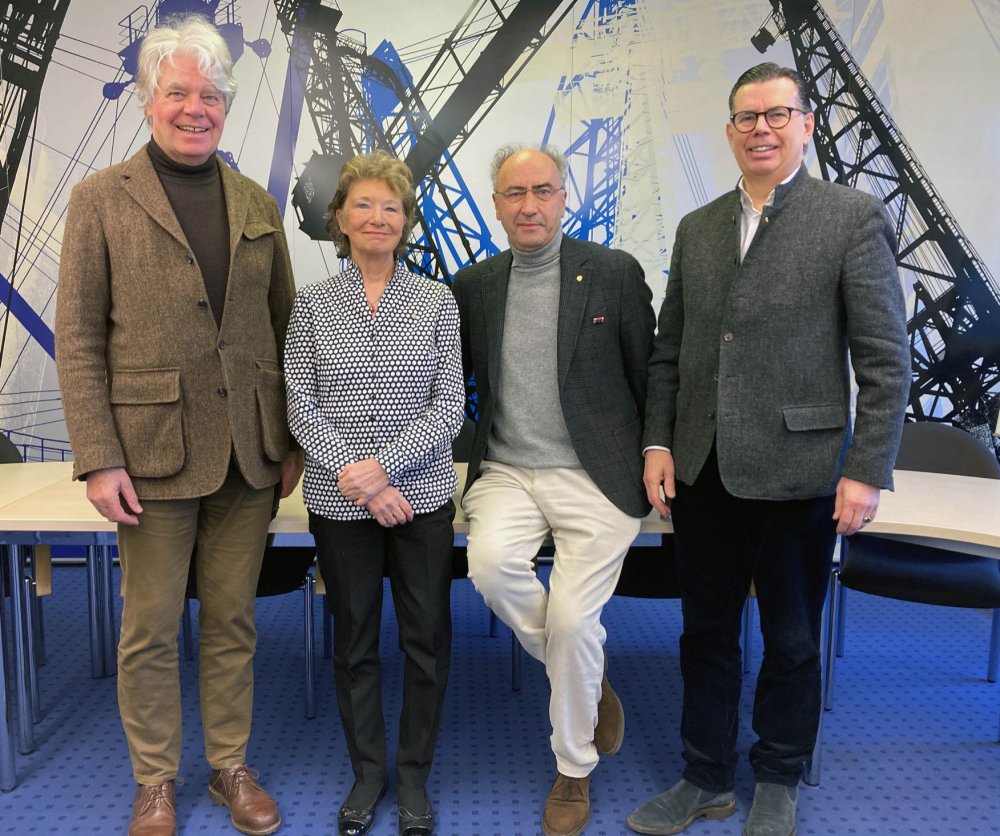 Vier Menschen, drei davon Männer, eine Frau, posieren für ein Gruppenbild und lächeln in die Kamera. Sie lehnen oder sitzen auf einem Tisch.