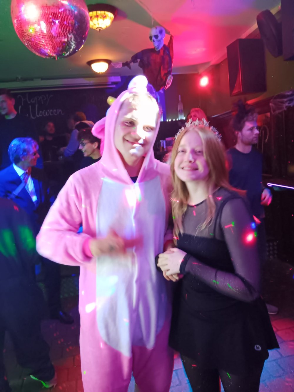 Auf einer Party stehen ein junger Mann im rosanen Kaninchen-Kostüm und eine junge Frau Arm in Arm, im Hintergrund sieht man feiernde Menschen und bunte Lichter.