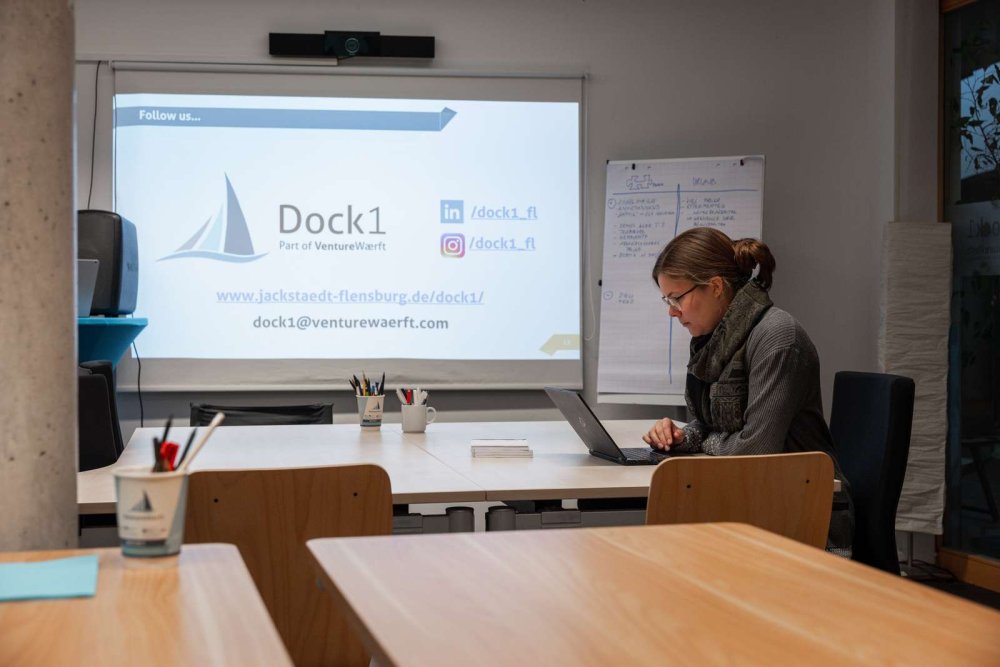 In einem Raum mit Schreibtischen sitzt eine junge Frau an einem Laptop und arbeitet, im HIntergrund sieht man an einer Leinwand eine Präsentation mit dem Logo des Dock1.
