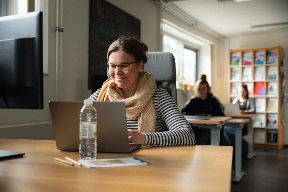 In einem Raum mit Schreibtischen sitzt eine junge Frau an einem Laptop und arbeitet, im Hintergrund sitzen auch an den anderen Tischen Menschen.