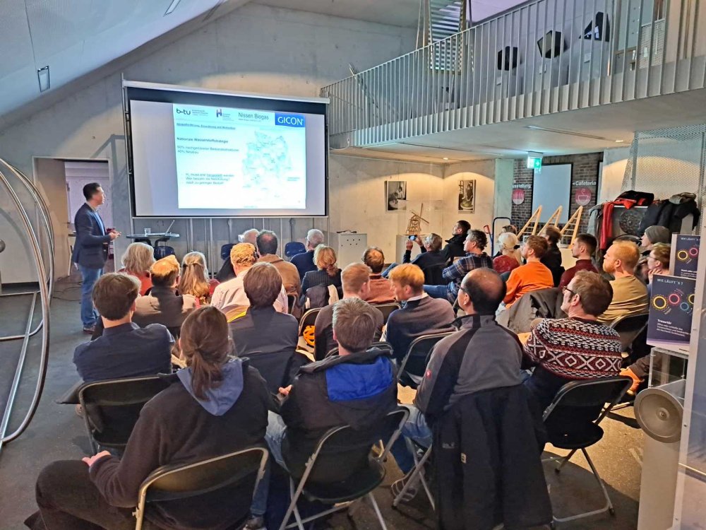 In einem Raum sitzen auf in Reihen aufgestellten Klappstühlen etwa 30 Menschen. Vorne steht ein Mann im Sakko und hält einen Vortrag mit einer PowerPoint-Präsentation, auf der verschiedene Logos und eine Deutschlandkarte zu sehen sind.
