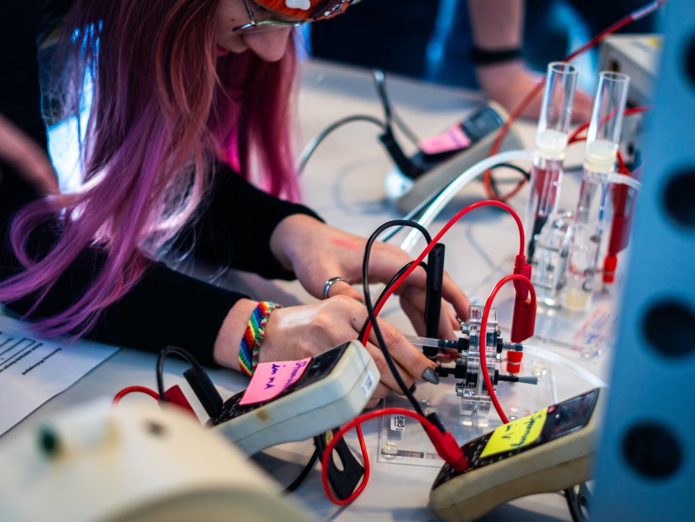 Eine Schülerin mit lilanen Haaren beugt sich über einen Wasserstoff-Versuchsaufbau mit vielen Schläuchen.